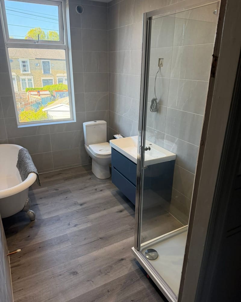 A bathroom finished by Graham, with grey tiles, a roll-top bath, a blue vanity unit and a glass shower enclosure