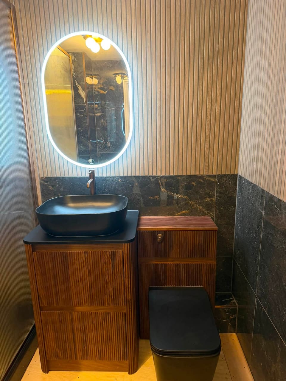 A finished bathroom by Graham: fluted walnut vanity unit with a black countertop basin, oval LED mirror and dark marble-effect tiles